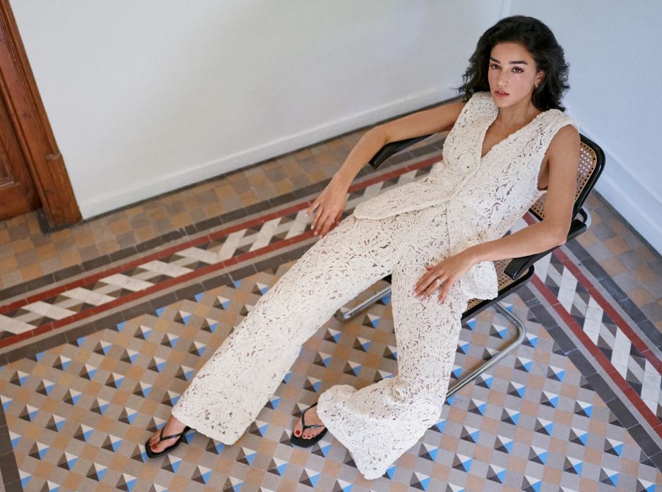 Mujer sentada en una silla, lleva un conjunto de blusa y pantalon color blanco tejido