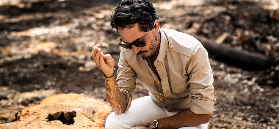 Hombre en cunclillas usando camisa beige, pantalon blanco y lentes para sol negros, KILIAN