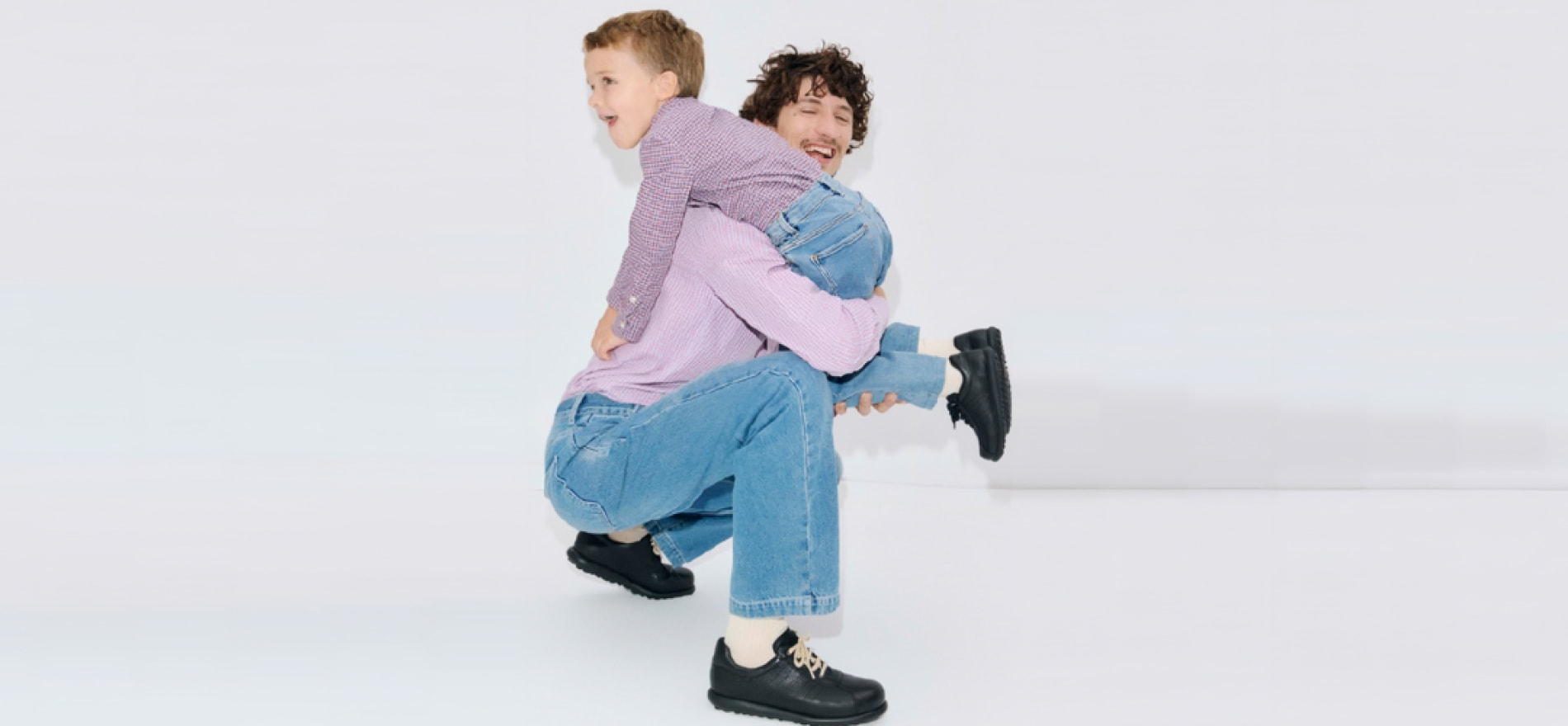 Padre sonriendo mientras carga a su hijo, ambos usando zapatos negros de la colecci&oacute;n de calzado infantil escolar, disponibles en El Palacio de Hierro
