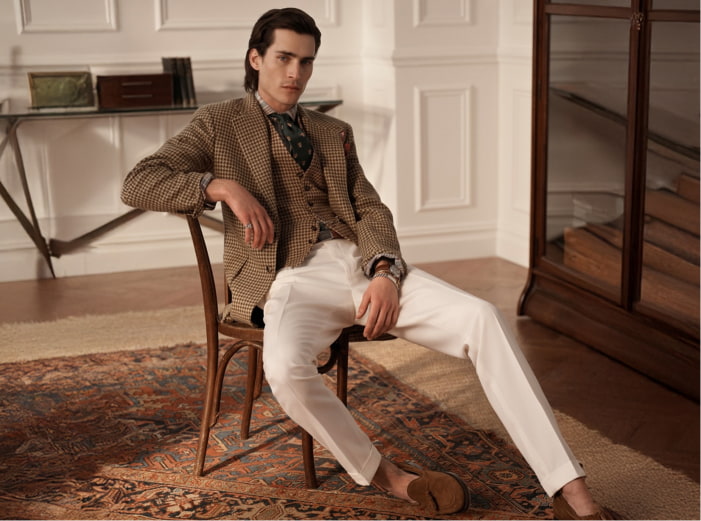 Hombre sentado en una silla lleva chaleco y saco color cafe y pantalon color crema