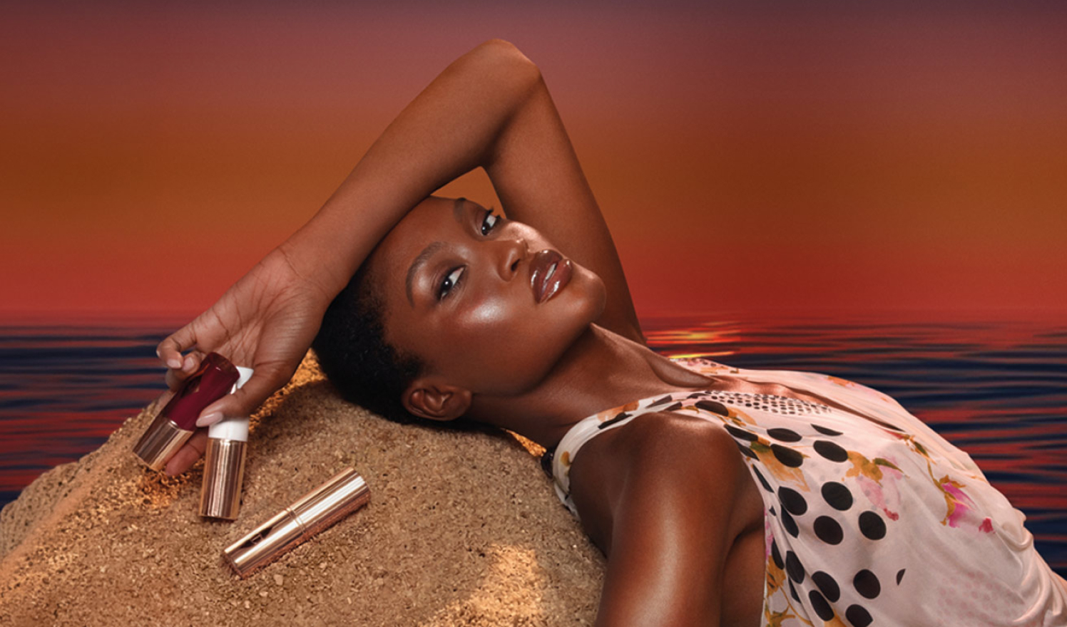 Modelo recostada en la arena al atardecer sosteniendo productos de maquillaje Charlotte Tilbury