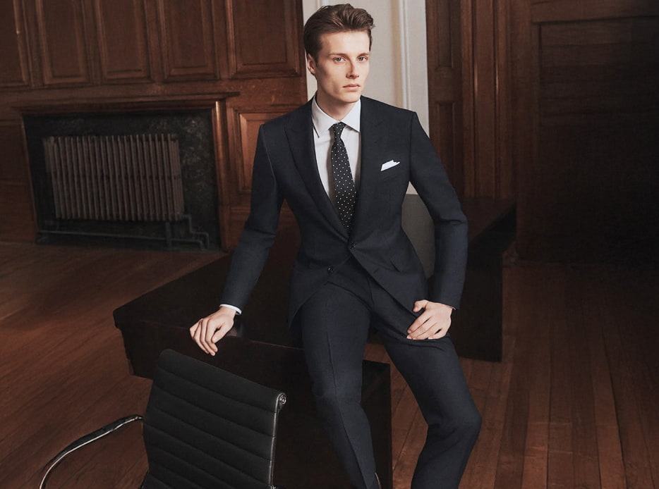 Hombre recargado en eun escritorio, lleva un traje color negro con camisa color blanca