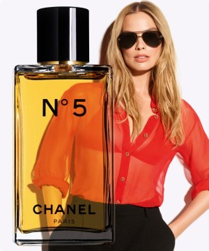 Imagen de una mujer con blusa transparente color anaranjada y pantalon negro, frente a ella un frasco de perfume CHANEL Nº5. banner menu