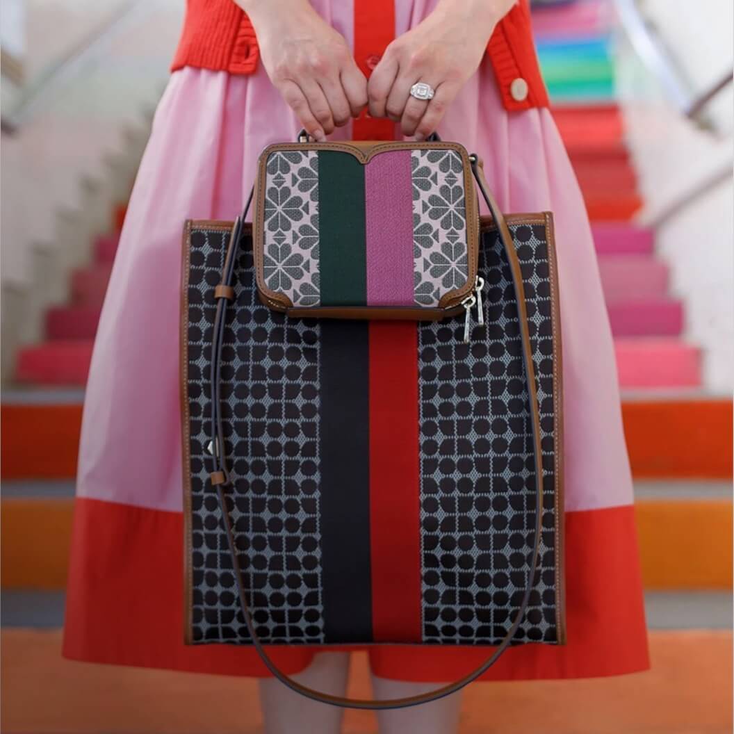 Imagen frontal de la marte inferior de una mujer, se observan sus piernas. Ella usa una falda larga, amplia, rosa con rojo, frente a esta sostiene un par de bolsas, una de mano y una más grande. Lista de marcas de Mujer
