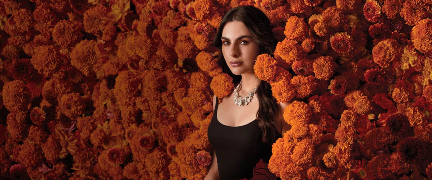 Imagen de una mujer con blusa negra entre una pared de flores de cempasuchil, Bien venido a noches palacio Día de Muertos
