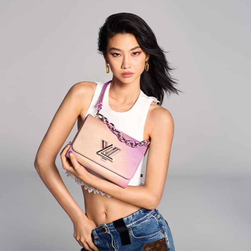mujer vistiendo playera corta blanca y pantalon de mezclilla azul, esta modelando un bolso de la marca LOUIS VUITTON, de colores difuminados morado con salmon, tiene una cadena grsande de color plateada con toques morados y en frente las letras LV de LOUIS VUITTON en color plateadas. LOUIS VUITTON