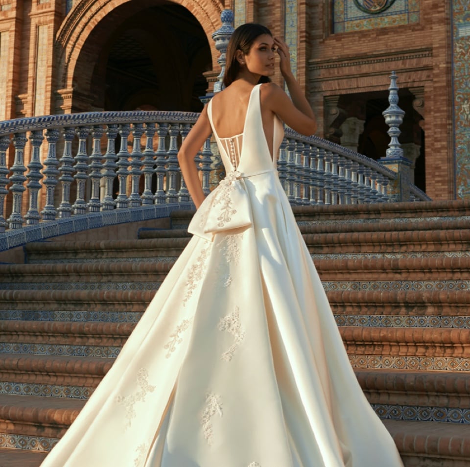 Imagen de mujer usando un vestido de novia blanco, satinado, con bordado de flores blancas, con escote debajo de las axilas y en la espalda, con tirantes, con moño grande en la espalda que termina en una cola. La mujer sube las escaleras de una construcción antigua y elegante, PRONOVIAS