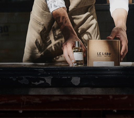 imagen hombre con mandil cafe enfrente de el , una botella trnsparente con tapa plateada de  fragancia. LE LABO