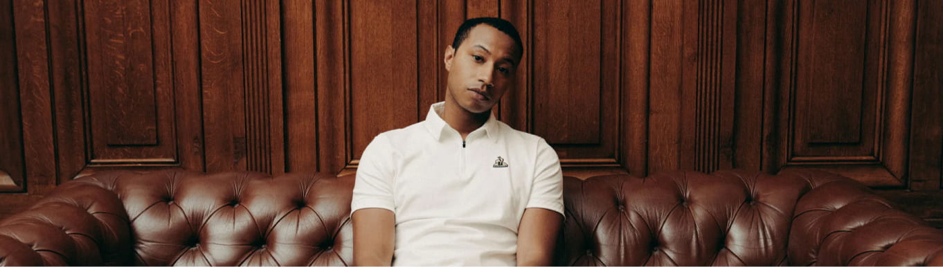 imagen de un hombre sentado en un sillon color cafe, lleva una playera color blanca con cuallo. LE COQ SPORTIF