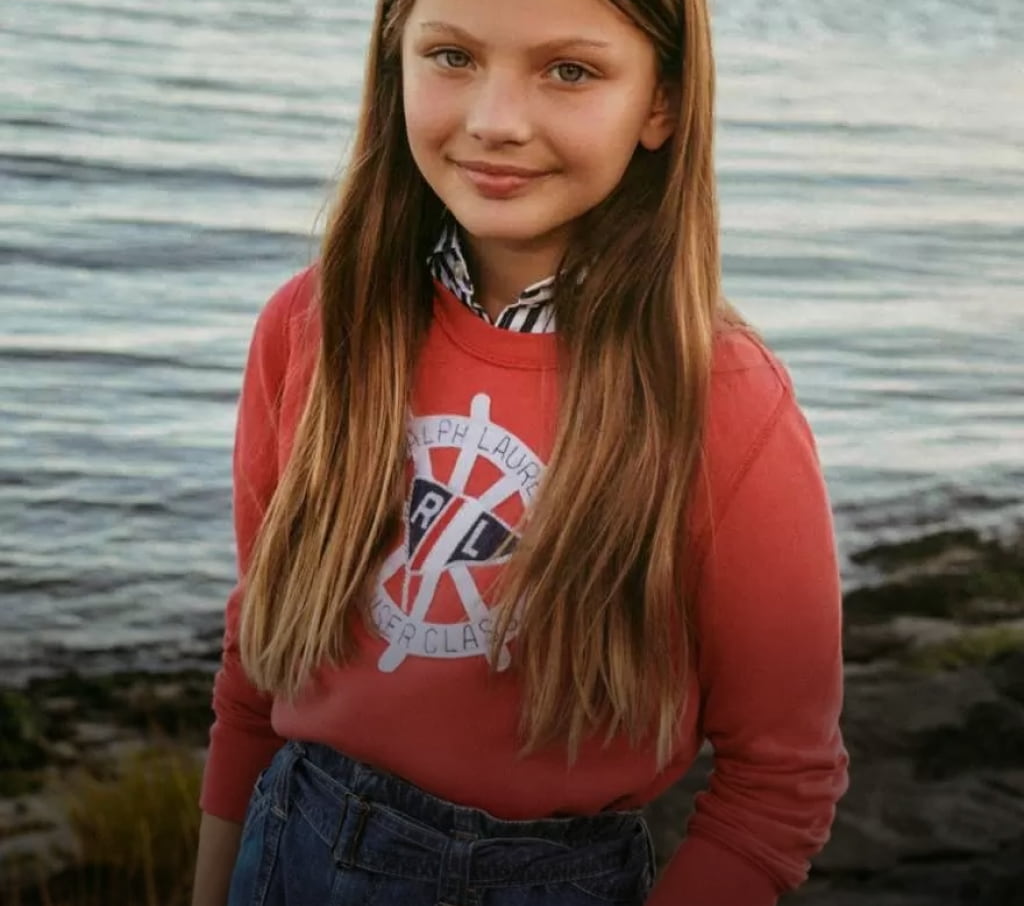 niñ con un sueter  rojo  de la marca POLO RALPH LAUREN, con arbustos de fondo y mar