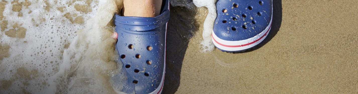 Imagen de unos pies a la oriya de una playa, usando unas sandalias de la marca CROCS color azul marino. CROCS