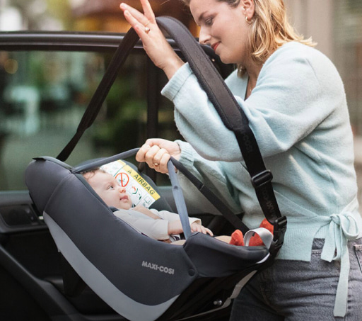 mujer con sueter azul y pantalon gris, metiendo un bebe, a su coche dentro de una silla para bebe para bebe de la marca MAXI COSI