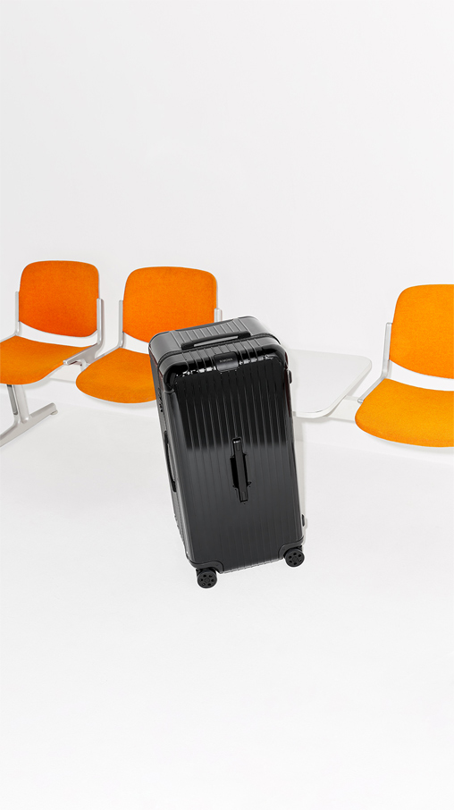 Imagen de una maleta rigida negra de la marca RIMOWA, en el fondo se ven sillas naranjas, RIMOWA