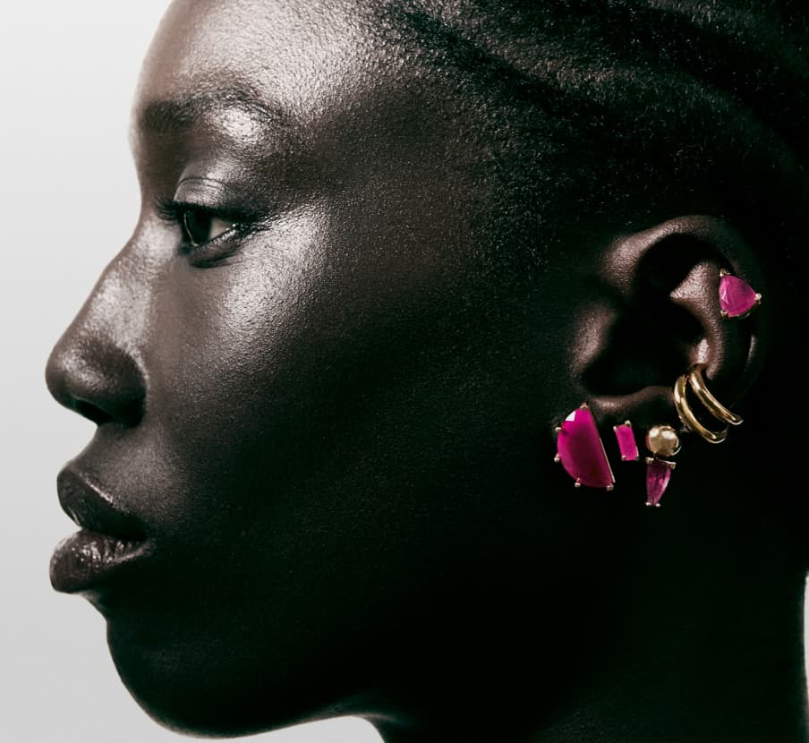 Imagen de la mitad izquierda del rostro de una mujer la cual lleva varios aretes dorados con piedras rosas. SUOT.