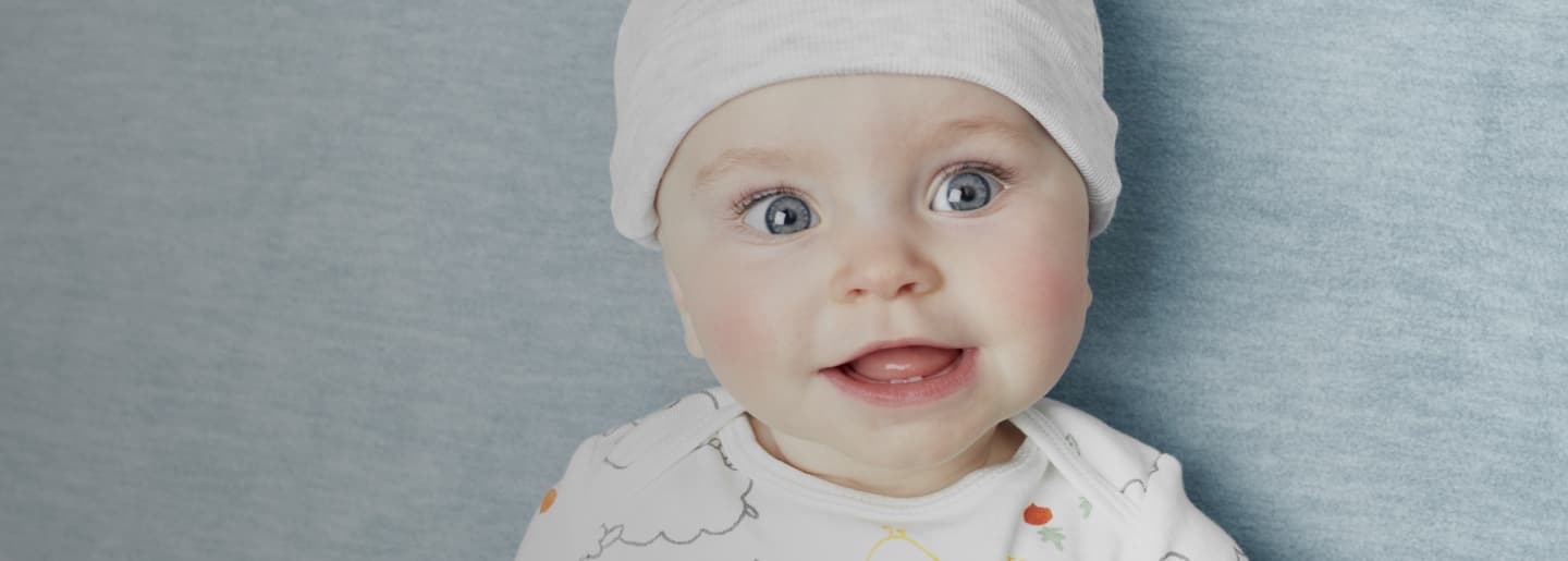 Rostro de bebé usando un gorro blanco y un pañalero blanco con estampado multicolor