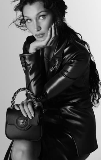 Mujer sentada, viste una chamarra negra de piel, una flada negra de latex y lleva un bolso negro de la marca VERSACE.  VERSACE