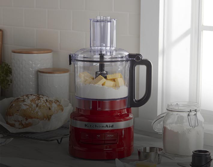 Imagen de una cocina en la cual se encuantra un procesador de alimentos de la marca KITCHEN AID, color rojo con su recipiente transparente. KITCHEN AID