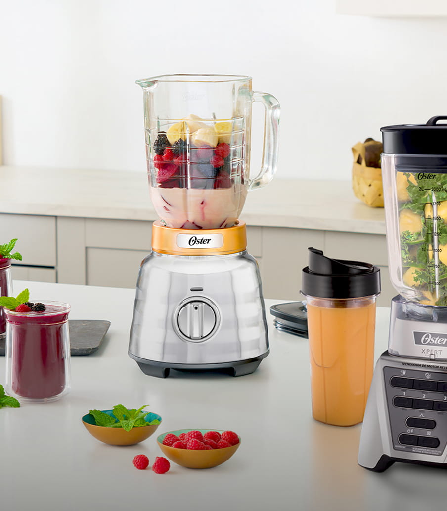 imagen de una cocina en ella se encuantran dos licuadoras de la marca OSTER sobre una barra, las licuadoras estas llenas de fruta listas para preparar jugos y junto a ellas dos vasos y un termo con jugo de frutas. OSTER