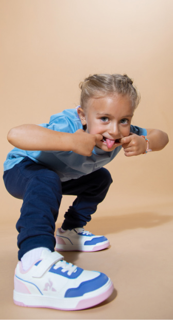 Imagen de una niña haciendo muecas, lleva una playeraazul cielo, pants azul rey y tenis blancos con toques rosa y azul cielo todo de la marca LE-COQ-SPORTIF