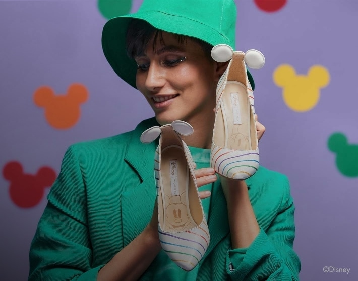 Mujer usando conjunto color verde de blusa, saco y gorro, en sus manos sostiene unas zapatillas de color blanco con con rayas de colores y en en tacon alto lleva unas orejas de Mickey mause , W CAPSULE
