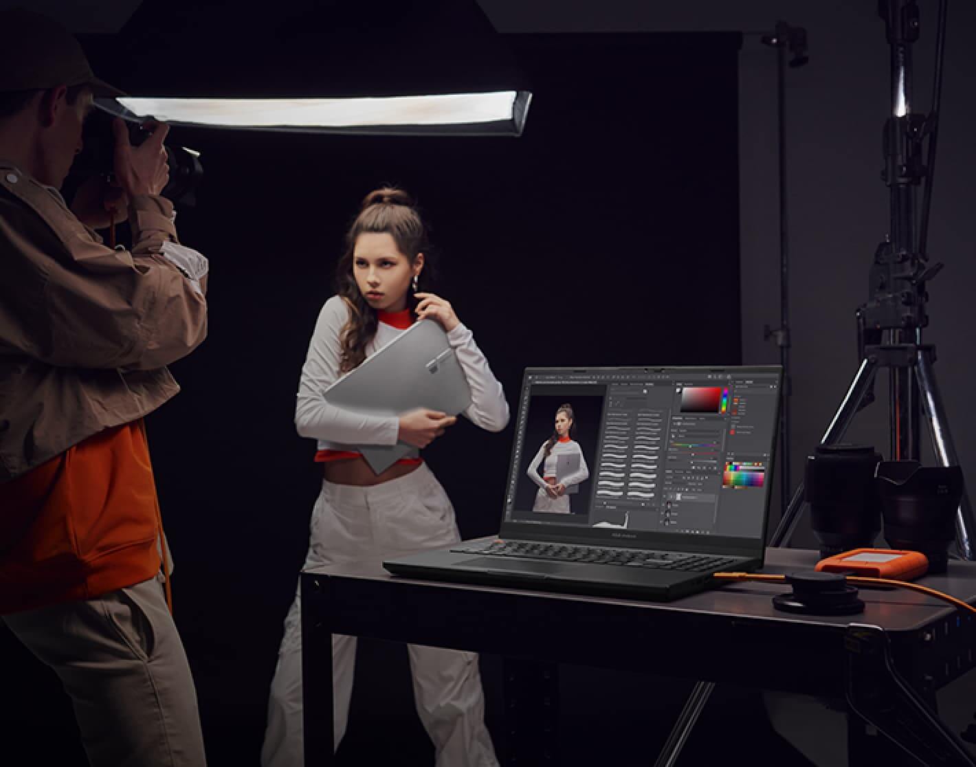 Imagen de un hombre y una mujer, ella esta modelando con una laptop plateada ASUS y el le esta tomando fotos, en un escritorio una laptop de la marca ASUS color negra en la cual estan editando las fotos. ASUS