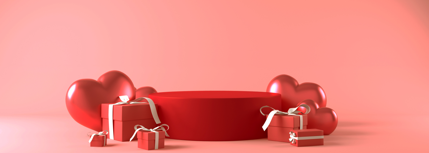 Imagen de cajas rojas de regalo de distintos tamaños, amarradas con liston blanco, atras de ellas tres coracones de diferentes tamaños. San Valentín