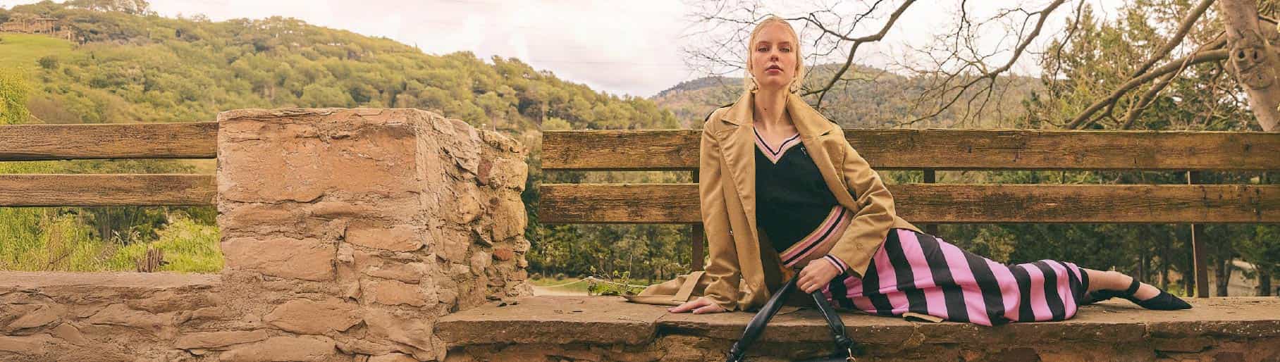 imagen de mujer acostada en banca de madera con chamarra cafe, blusa negra y falda  negra con rosa la marca LOLA CASADEMUNT 