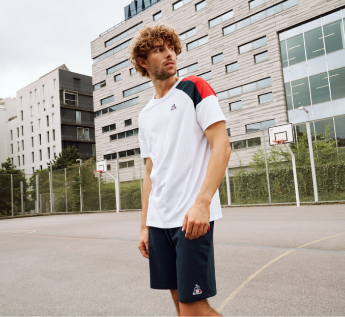 Imagen de un hombre en una cancha de basquetbol, lleva playera blanca con raya azul y roja en hombros y short color azul marino con logo de una gallo de la marca LE-COQ-SPORTIF