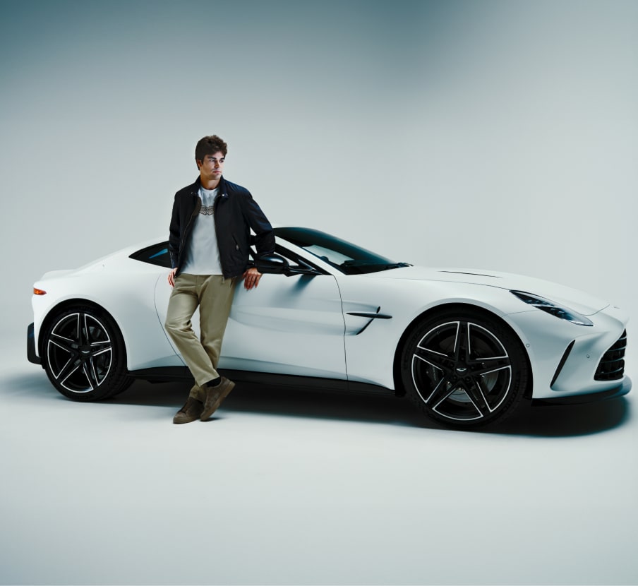 Imagen de un hombre usando una chamarra azul, playera blanca, pantalon y tenis cafes junto a automivil blanco de la marca, HUGO BOSS