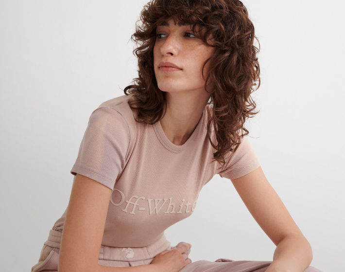 Mujer sentada, usando un conjunto de playera y pants de color salmon claro, la playera tien el nombre de la marca OFF WHITE en color blanco. OFF WHITE