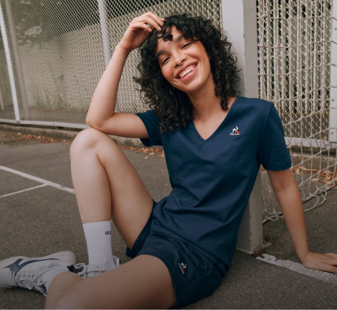 Imagen de una mujer sentada en una cancha, lleva un conjunto de playera y short azul marino con logo de una gallo de la marca LE-COQ-SPORTIF
