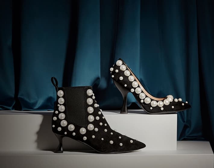Imagen de dos zapatos de modelos distintos, uno es un zapato de tacón negro con perlas blancas de fantasía de adorno, y el otro es un botín de tacón bajo con perlas blancas de fantasía de adorno. MOSCHINO
