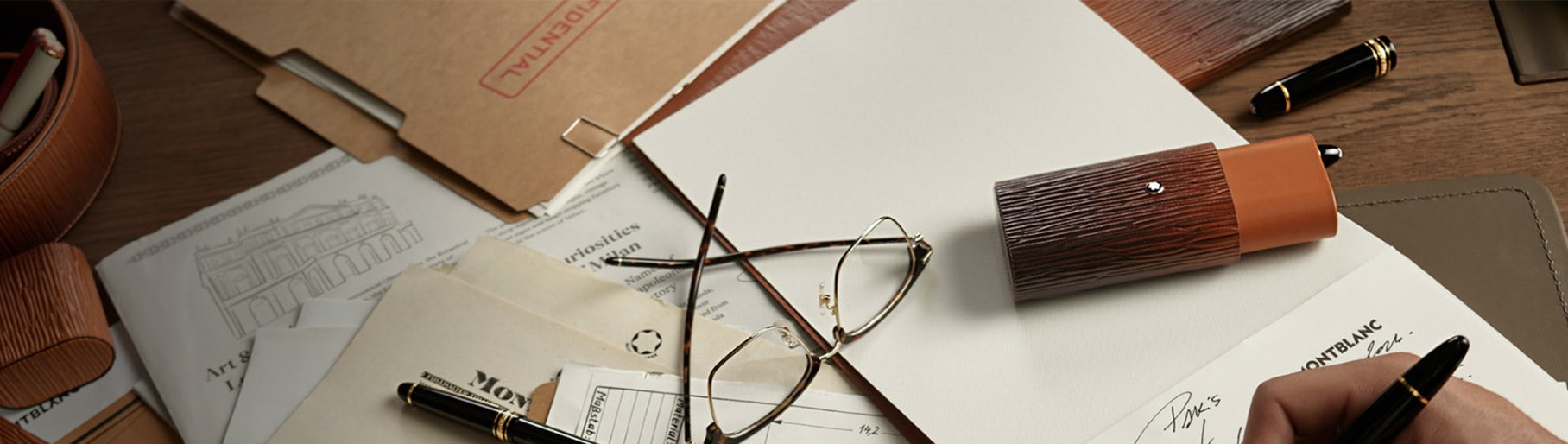 Imagen de una mesa, sobre ella unos lentes y varios papeles, una mano usando una pluma negra de la marca MONTBLANC