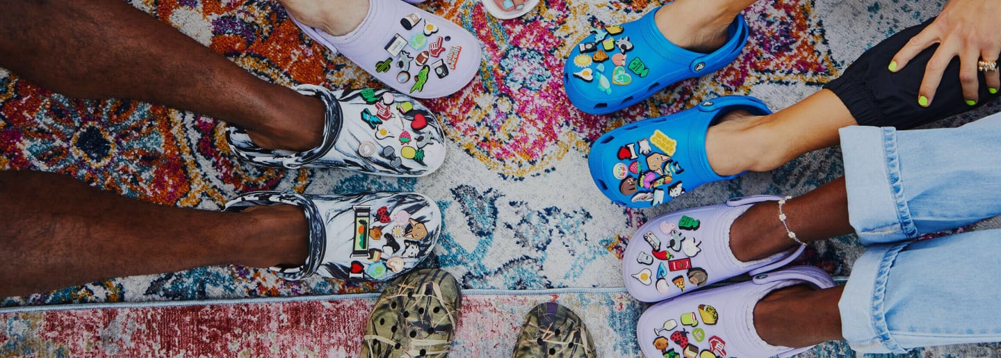Imagen de sinco pares de pies los cuales llevan sandalias de la marca CROCS, de diferentes modelos y colores para hombre y mujer. CROCS