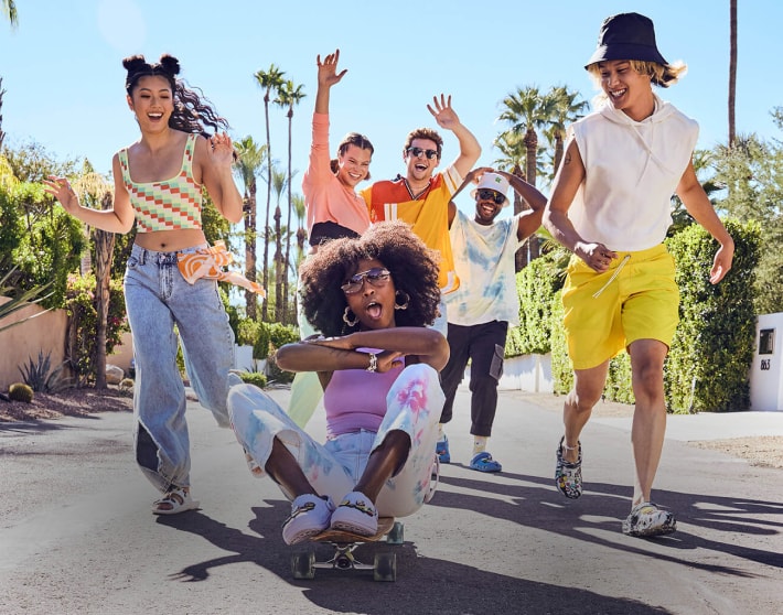 imagen de seis jovenes jugando en la calle sonriendo, todos llevan sandalias de la marca CROCS de siferentes modelos, al frente una mujer sentada en una patineta usando CROCS de color blancos con pines. CROCS