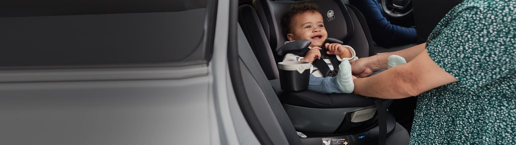 Imagen de un bebé con ropa blanca y azul, sentado en sillita para auto movil, lo esta asegurndo una mujer con vestido negro con flores verdes, Articulos para bebe