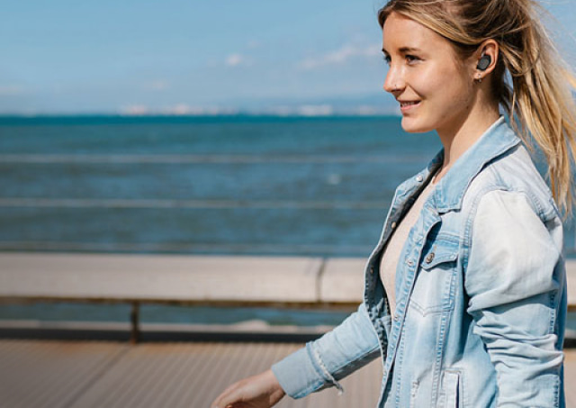 Imagen de mujer caminando que lleva audifono inalambrico gris de la marca BELKIN y chamarra de mezclilla con el mar de fondo