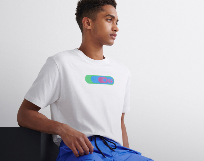 Hombre sentado, usando una playera de la marca OFF WHITE color blanca con estampado de color verde y azul y la palafra OFF en color blanco en la parte de enmedio y un pants color azul. OFF-WHITE