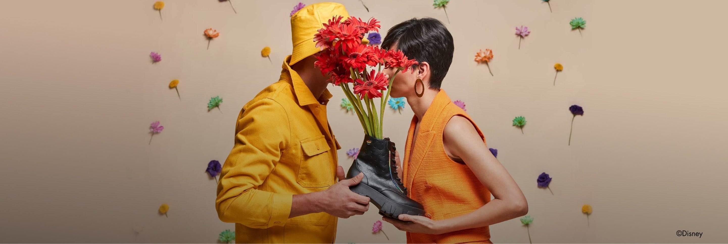 Imagen de un hombre y una mujer viendose cara a cara y sosteniendo una bota negra con unas margaritas en su interior, el lleva un impermeable amarillo, ella una blusa color naranja, W CAPSULE