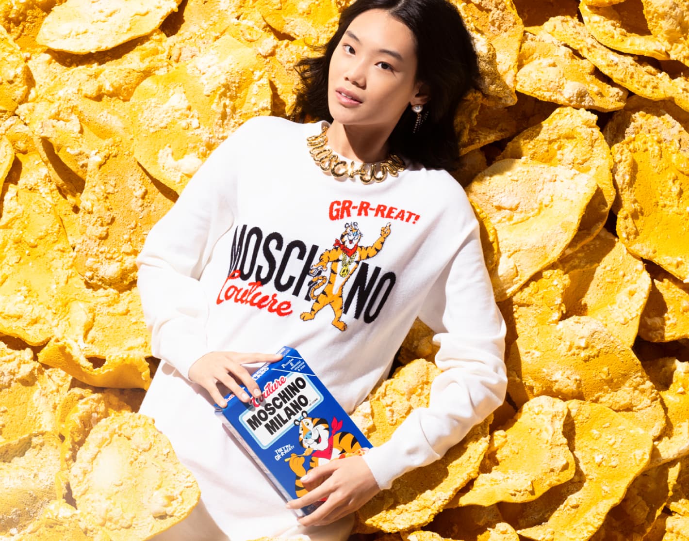 Imagen de una mujer acostada sobre ojuelas de maiz, lleva una sudadera blanca con la marca MOSCHINO y sistine una caja de ceral. MOSCHINO