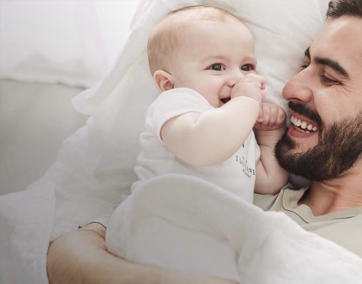 Bebé usando un pañalero blanco, recostado en una cama siendo abrazado por su padre. Ambos sonríen. CARTER'S