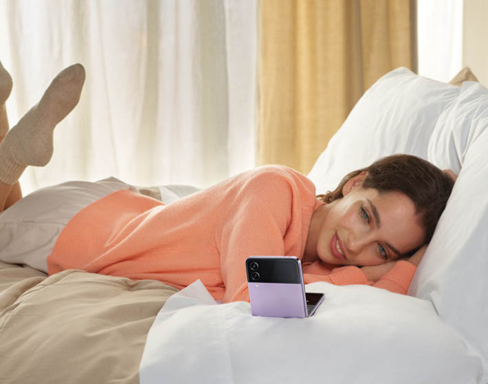 Imagen de una mujer acostada en una cama llevanmdo sueter coral y pantalon crema mientras ve su celular SAMSUNG