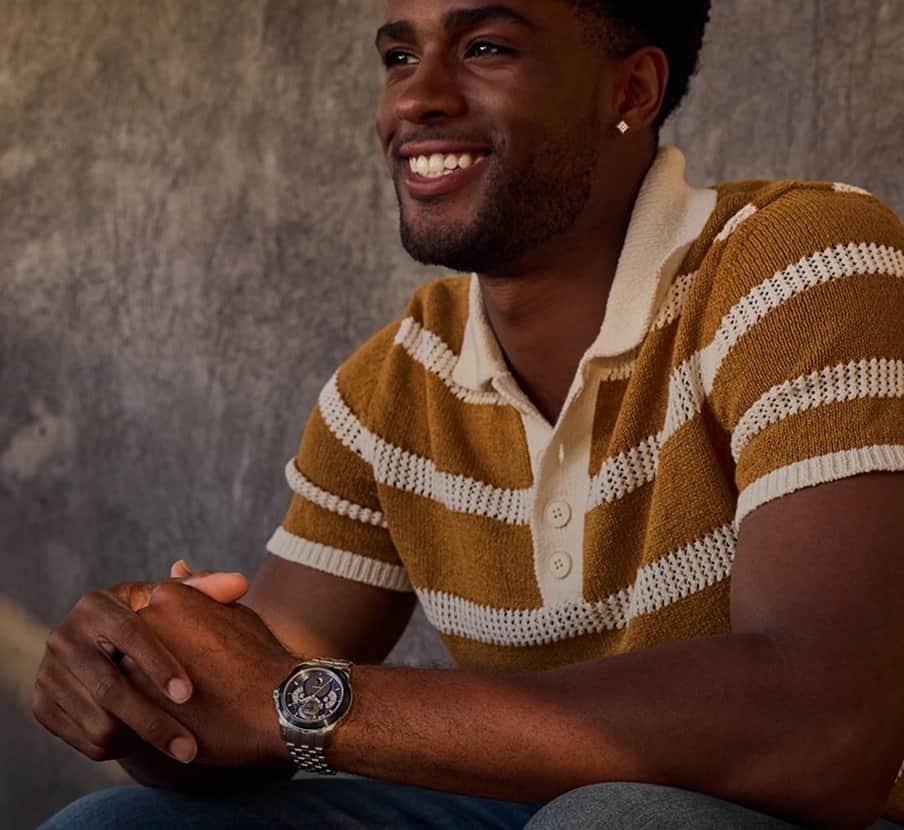 Imagen de hombre usando playera tipo polo tejida, a rayas blancas con cafes, lleva en mano izquierda un reloj plateado con caratula azul. CITIZEN
