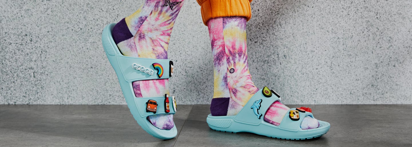 Imagen de unos pies usando calcetas blancas con muchos colores y unas sandalias de la marca CROCS, color azul claro con pines. CROCS