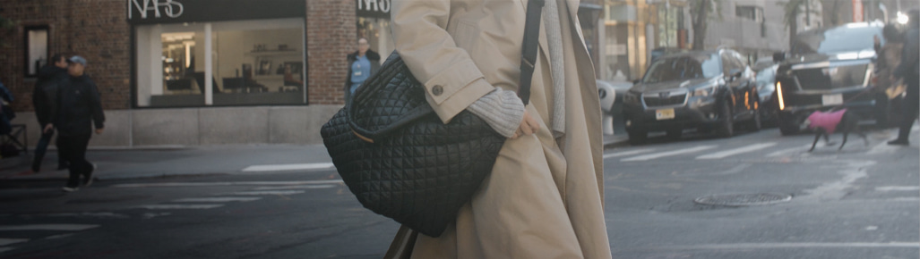 imagen de una mujer cruzando la calle. con automoviles y edificios de fondo, ella lleva una gabardina cafe y lleva un bolso negro de un hombro de la marca RAZER