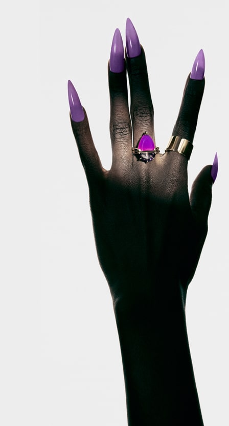 Imagen de la mano de una mujer con uñas postizas moradas y en sus dedos un anillo plateado con piedra color morada. SUOT