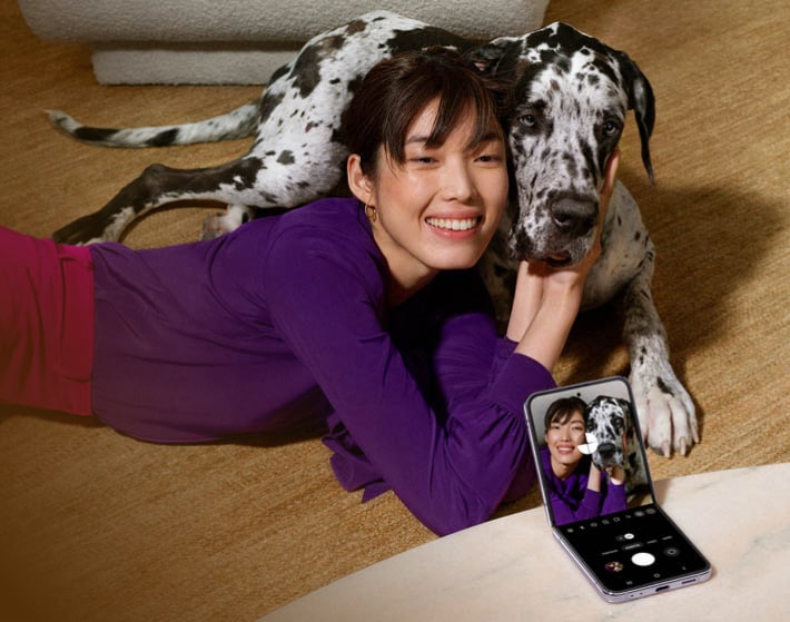 Imagen de una mujer acostada en el suelo con su perro a un lado llevando un sueter morado tomando se una foto con su celular SAMSUNG
