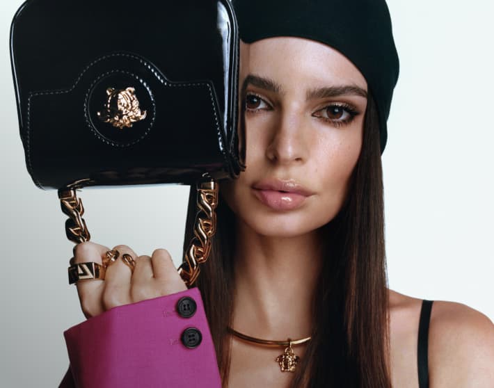 Una mujer sosteniendo un bolso de la marca VERSACE color negro con asa en forma de cadena dorada. VERSACE