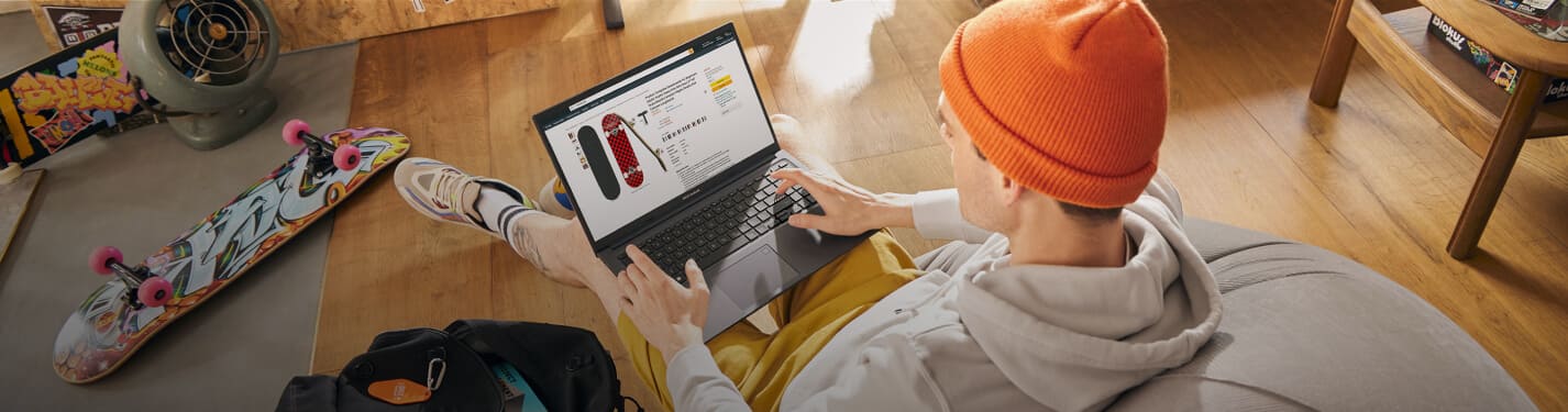 Imagen de un hombre sentado en sillon pop, viste una sudadera gris, pantalon amarillo y gorro color rojo, esta usando una laptop de la maraca ASUS, color negra en la cual esta viendo una pagina de compras. ASUS