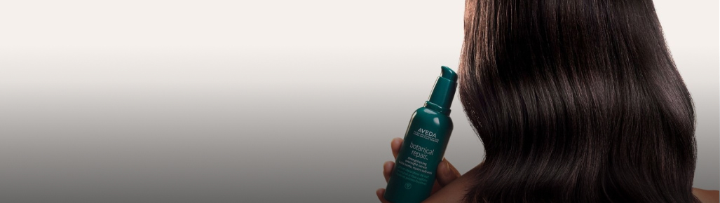 mujer de cabello castaño largo de espalda trae una botella verde de tratamiento botanical repair de la marca AVEDA